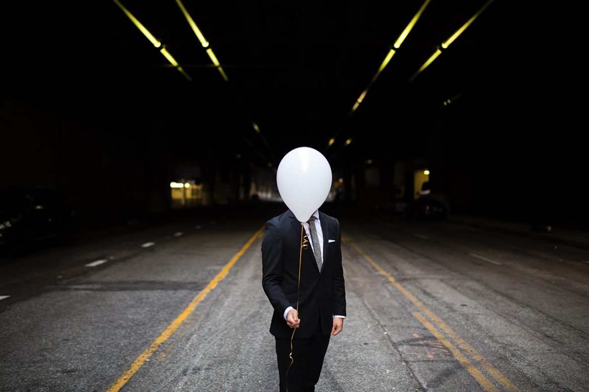 Photo of a man in a suite holding a white balloon standing in the middle of the road.